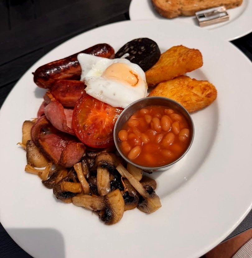 A full cooked breakfast: sausages, bacon, black pudding, a fried egg, hash browns, grilled tomato, mushrooms, beans in a pot, with toast on the side